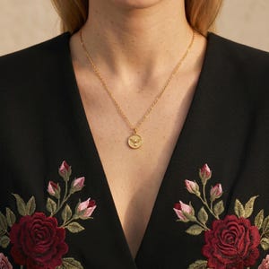 Puede incluir: Un collar dorado con un colgante redondo con un dise&ntilde;o de p&aacute;jaro. El collar se lleva sobre una blusa negra con bordados florales rojos y rosas. El colgante cuelga en el escote, creando un look sencillo pero elegante.