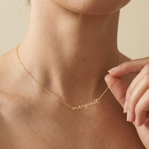 May include: A gold-toned necklace featuring a delicate chain and a central pendant formed by the name "margaret" written in a cursive, lowercase script font. The metal has a polished finish that reflects light, creating a bright, metallic appearance.