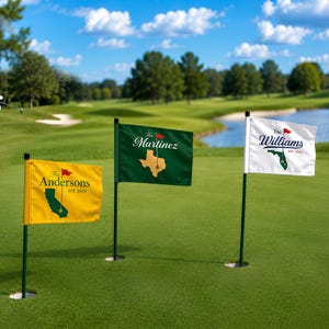Op de afbeelding: Drie gepersonaliseerde golfflaggen op een groene golfbaan. De vlaggen zijn geel, groen en wit, elk met een andere naam en een staatsomtrek. Op de achtergrond zwaait een golfer met een golfclub.