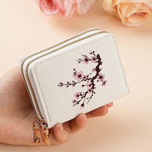 May include: A white, textured faux leather wallet featuring a double zip closure with gold-toned hardware. The front of the wallet is decorated with a printed illustration of a dark brown tree branch adorned with small, light pink cherry blossoms.