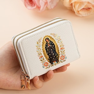 May include: A white, textured faux leather wallet featuring a double zipper closure with gold-toned pulls. The front displays a colorful, detailed illustration of the Virgin of Guadalupe surrounded by a floral wreath and golden rays of light.