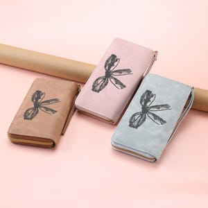 May include: Three rectangular zip-around wallets in tan, light pink, and light blue. Each wallet features a black graphic print of a lace-style bow on the front surface and includes a matching wrist strap attached to the side with a gold-tone zipper.