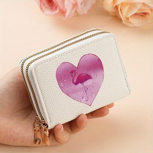Peut inclure: Un portefeuille rectangulaire blanc à texture grainée, doté d'une double fermeture éclair avec des tirettes dorées. Le devant du portefeuille présente un grand cœur rose de style aquarelle avec la silhouette d'un flamant rose.