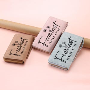 May include: Three rectangular zippered wallets in tan, dusty pink, and light blue. Each wallet features a black graphic print of two small flowers and the text "Fear not ISAIAH 41:10" in a cursive script. The wallets have gold-toned zippers.