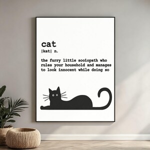 May include: A white rectangular wall art print featuring black text that defines a cat as a furry little sociopath who rules your household and manages to look innocent while doing so, accompanied by a black silhouette of a lying cat.