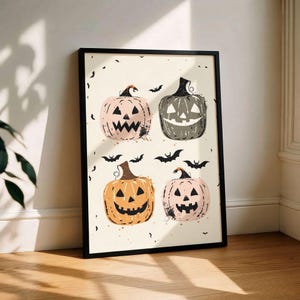 May include: A framed wall art print featuring four stylized jack-o'-lanterns in shades of light pink, dark gray, orange, and pale peach. The pumpkins have carved faces and are surrounded by small black bat silhouettes on a cream background.