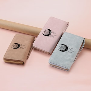 May include: Three pastel-colored wallets with a celestial design. The wallets are in shades of tan, pink, and blue-green. Each wallet features a black crescent moon with a lotus flower and dangling beads. The wallets have a zipper closure and a wrist strap.
