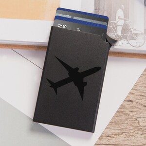 May include: A black cardholder with a silhouette of an airplane on the front. Several cards are partially visible inside. The cardholder is rectangular and appears to be made of metal or a similar material.