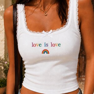 May include: A white ribbed tank top featuring a square neckline with delicate white lace trim and a small white bow at the center. The chest displays the embroidered text "love is love" in multicolored letters above a small rainbow graphic.