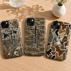May include: Three smartphone cases featuring camouflage patterns. The left case displays a collage of a deer, fish, turkey, and tractor. The center case features three black crosses. The right case shows a mallard duck in flight over a brown camo print.