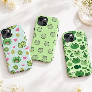 May include: Three smartphone cases featuring frog-themed designs. The left case is light blue with frogs, lily pads, and pink hearts. The middle case is light green with a repeating frog pattern. The right case is green with frogs and foliage.