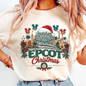 May include: A cream-colored t-shirt featuring a graphic of Epcot with a Santa hat, a monorail, and holiday decorations. The text reads EPCOT Christmas AROUND THE WORLD, surrounded by small Mickey Mouse ear icons and festive greenery.