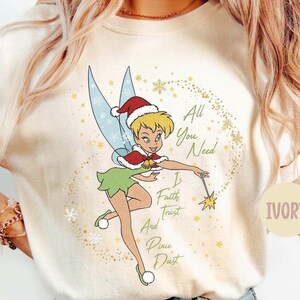May include: An ivory t-shirt featuring a graphic of a fairy wearing a red and white Santa hat and cape. The text reads All You Need Is Faith Trust And Pixie Dust, surrounded by gold sparkles and snowflakes. The shirt has short sleeves and a crew neck.