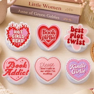 May include: Six heart-shaped phone grips featuring various designs and text: Hot Girls Read, Book girlie, best Plot twist, Book Addict, Please don't talk to me while I'm reading, and Kindle Girlie, all in shades of pink, red, and white.