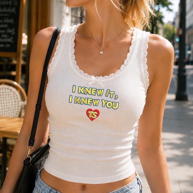 Puede incluir: Una camiseta de tirantes blanca acanalada con ribete de encaje en el escote y un peque&ntilde;o lazo en el centro. El frente presenta el texto I KNEW IT, I KNEW YOU en amarillo con bordes azules, sobre un coraz&oacute;n rojo con las letras amarillas TS.