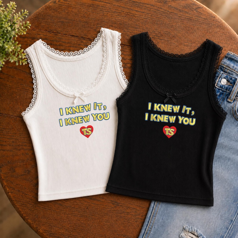 Puede incluir: Una camiseta de tirantes blanca y otra negra de canal&eacute;, ambas con escote redondo ribeteado en encaje y un peque&ntilde;o lazo central. Las prendas muestran el texto amarillo I KNEW IT, I KNEW YOU sobre un coraz&oacute;n rojo con las letras amarillas TS.