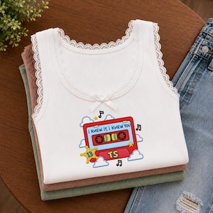 May include: A white ribbed tank top with lace trim along the neckline and a small bow. The front features an embroidered red cassette tape graphic with the text "I KNEW IT, I KNEW YOU" and "TS" inside, surrounded by clouds, stars, and musical notes.