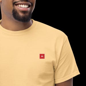 Pode incluir: Uma t-shirt de manga curta em amarelo claro com um pequeno emblema quadrado vermelho no peito. O emblema contém uma ilustração gráfica branca de um pico de montanha sob um sol circular, centrado no tecido da peça de vestuário.