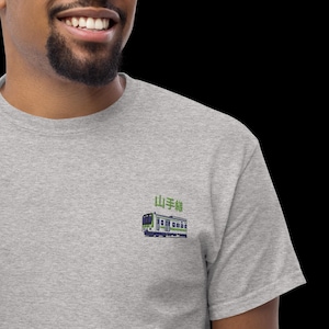 Pode incluir: Uma t-shirt cinzenta mesclada com um gráfico bordado de um comboio suburbano com detalhes em verde e branco. Por cima do comboio, os caracteres japoneses 山手線 estão bordados em fio verde. O design situa-se no lado esquerdo do peito.