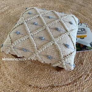 May include: A square, beige decorative pillow with a diamond pattern and small blue tufts. The pillow is made of a textured material and sits on a woven jute rug. A book with geometric designs is partially visible.