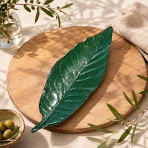 Può includere: Un vassoio in ceramica verde scuro lucido a forma di foglia allungata con venature visibili sulla superficie. Il vassoio presenta una finitura liscia e riflettente e una punta affusolata, che imita la forma naturale di una foglia botanica.