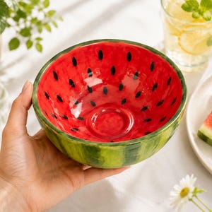 May include: A round ceramic bowl painted to resemble a watermelon, featuring a bright red interior with black seed-like markings and a green exterior with darker green vertical stripes, held in a palm to show its size and circular shape.