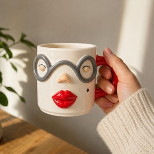 Può includere: Una tazza in ceramica con un design a volto. La tazza ha una base color crema con una montatura di occhiali grigia, un naso piccolo e labbra rosse. Il manico è rosso. Un piccolo punto nero è sul lato destro del volto.