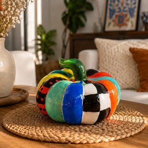 Op de afbeelding: Een decoratieve keramische pompoen met een glanzende afwerking en levendige, veelkleurige verticale segmenten. De segmenten bevatten patronen van zwart-witte blokjes, effen blauw, feloranje, turquoise en rood met zwarte stippen.