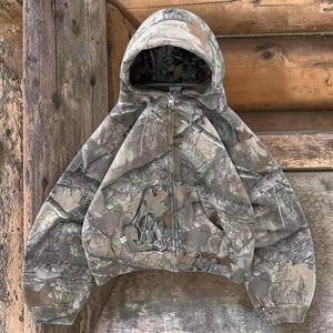 Könnte beinhalten: Ein Kapuzen-Sweatshirt mit Reißverschluss und Camouflage-Muster in Braun-, Grün- und Grautönen. Das Sweatshirt hat eine Vordertasche und einen durchgehenden Reißverschluss. Die Kapuze ist mit einem ähnlichen Camouflage-Muster gefüttert.