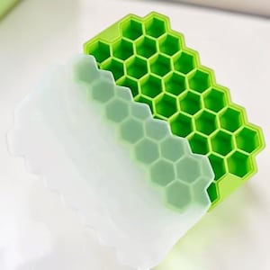 May include: A green silicone ice cube tray with a honeycomb design. The tray is partially covered by a white, flexible lid. The honeycomb cells are visible through the lid. The tray is designed for making ice cubes.