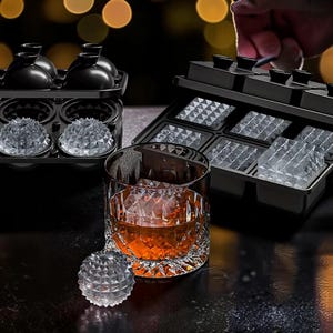 Può includere: Un bicchiere da whisky pieno di liquido ambrato e grandi cubetti di ghiaccio trasparenti. Sono visibili due vassoi per cubetti di ghiaccio neri, uno con stampi per ghiaccio sferici e l'altro con stampi quadrati. Un singolo cubetto di ghiaccio sferico è appoggiato davanti al bicchiere.