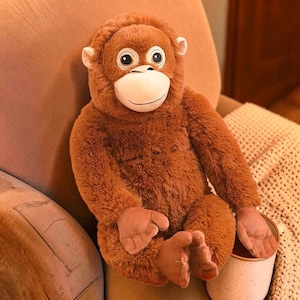 Puede incluir: Un peluche de orangután con pelaje marrón, cara blanca y ojos grandes y expresivos. El animal de peluche está sentado en una silla, con los brazos y las piernas extendidos. Sostiene una taza blanca.