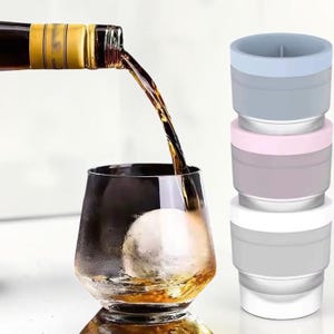 May include: A glass of amber liquid with a large ice sphere is being filled from a bottle. Stacked next to the glass are three collapsible silicone containers in pastel colors: blue, pink, and white. The glass has a unique, angular shape.