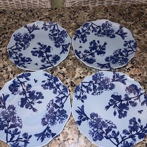 May include: A set of four light blue plates with a scalloped edge, each featuring a dark blue floral pattern of cherry blossoms and branches. The plates are round with a smooth surface and a consistent botanical design across all four pieces.