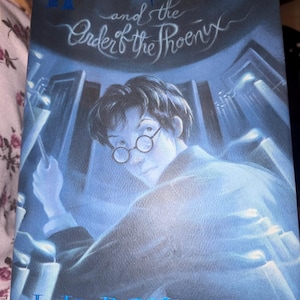May include: A blue-toned book cover for Harry Potter and the Order of the Phoenix by J.K. Rowling. The illustration features a young person with dark hair and round glasses holding a wand, surrounded by glowing light and tall structures.