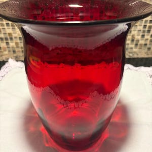 May include: A deep ruby red glass vase with a wide, flared rim and a smooth, rounded body. The glass features a translucent quality that allows light to pass through, highlighting the vibrant crimson color and the subtle, curved silhouette.