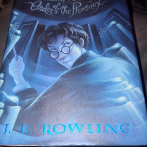 May include: A hardcover book titled Harry Potter and the Order of the Phoenix by J.K. Rowling. The blue cover features an illustration of a young person with dark hair and round glasses holding a wand amidst glowing candles.