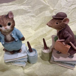 May include: Two ceramic figurines featuring a brown tabby kitten in a blue jacket standing on a stack of books titled The Tale of Tom Kitten and a brown mouse in a brown coat and hat holding a briefcase standing on books titled The Tale of Johnny Town-Mouse.