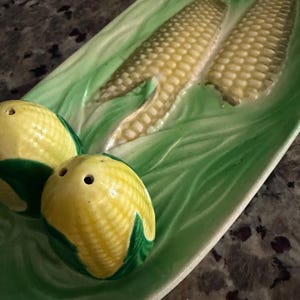 May include: A ceramic corn-shaped salt and pepper shaker set. The set includes two corn-shaped shakers and a serving dish with embossed corn cobs and green leaves. The shakers are yellow with green husks, and the dish is green.