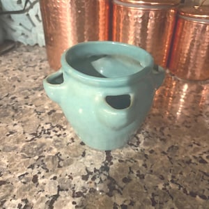 May include: A small, turquoise ceramic pot with two side handles. The pot has a rounded shape and a smooth, glossy finish. Copper canisters with lids are in the background, suggesting a kitchen or home decor theme.