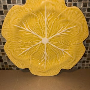 May include: A yellow ceramic plate shaped like a large cabbage leaf with a scalloped edge. The surface features a raised, textured pattern of white veins radiating from the center, mimicking the natural appearance of a leafy vegetable.