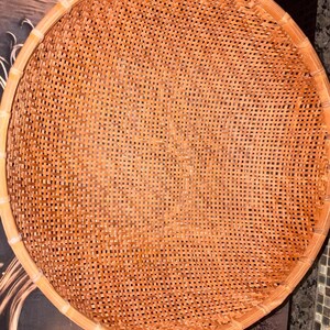 May include: A round, shallow basket featuring a woven lattice pattern made from thin, light brown strips. The basket has a thick, reinforced circular rim constructed from a smooth, lighter-colored material that frames the intricate, open-weave base.