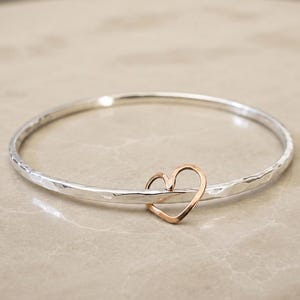 Peut inclure: Un bracelet jonc argenté avec une texture martelée et un charm en forme de cœur en or rose. Le cœur est attaché au bracelet, créant un design simple mais élégant. Le bracelet est présenté sur une surface neutre.