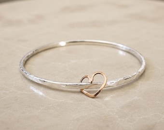 May include: A silver bangle bracelet with a hammered texture and a rose gold heart charm. The heart is attached to the bracelet, creating a simple yet elegant design. The bracelet is shown on a neutral surface.