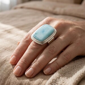May include: A silver ring featuring a large, rectangular, pale blue gemstone. The ring is displayed on a hand, with the gemstone's colour contrasting against the skin tone. The ring's design is simple, with a polished silver band.