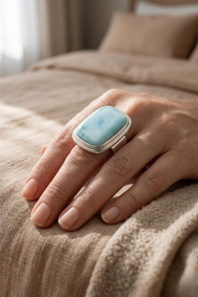 May include: A silver ring featuring a large, rectangular, pale blue gemstone. The ring is displayed on a hand, with the gemstone's colour contrasting against the skin tone. The ring's design is simple, with a polished silver band.