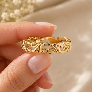 Puede incluir: Un anillo de oro con un intrincado diseño calado continuo de patrones de filigrana en espiral. El metal tiene un acabado pulido y reflectante que capta la luz, resaltando las curvas y bucles detallados que forman la estructura de la banda.