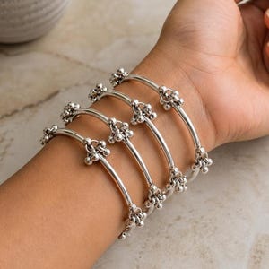 Puede incluir: Un conjunto de cuatro brazaletes plateados. Cada brazalete presenta un diseño curvo y liso, adornado con un grupo de pequeñas cuentas redondas en el centro y en los extremos. Los brazaletes se lucen en una muñeca.