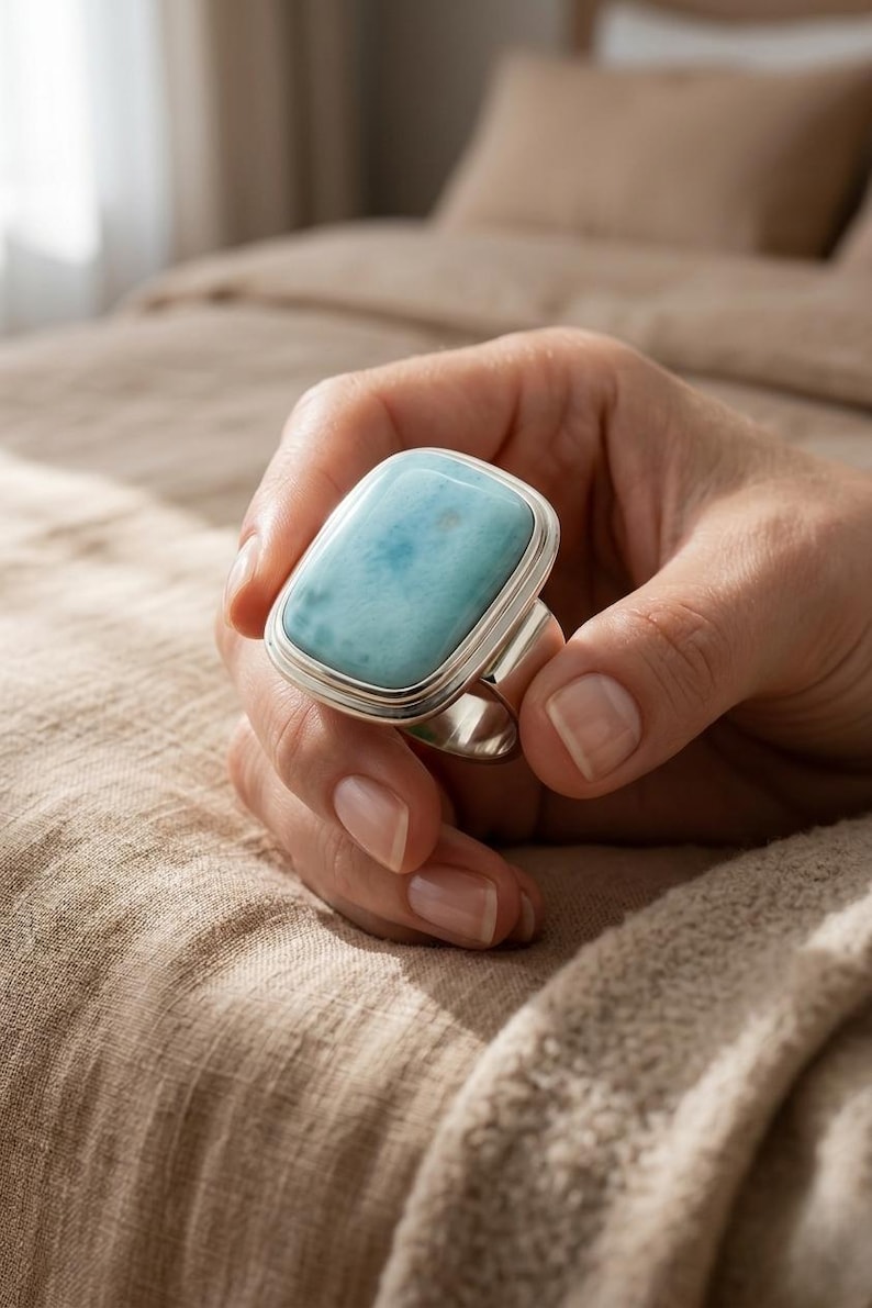 May include: A silver ring with a rectangular, pale blue gemstone. The ring is held in a hand, highlighting the gemstone's colour and shape. The gemstone has a soft, cloudy appearance. The background is a neutral, warm tone.