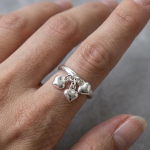Könnte beinhalten: Ein silberner Ring mit drei herzförmigen Anhängern. Der Ring wird an einem Finger getragen und zeigt das zarte Design und die polierte Oberfläche der Herzanhänger. Der Ring hat ein schlichtes Band, das die Herzen hervorhebt.